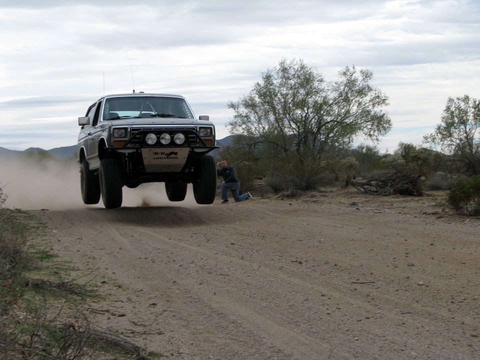 1980 Ford Bronco jumping a dirt road in a desert landscape with a photographer kneeling to the side