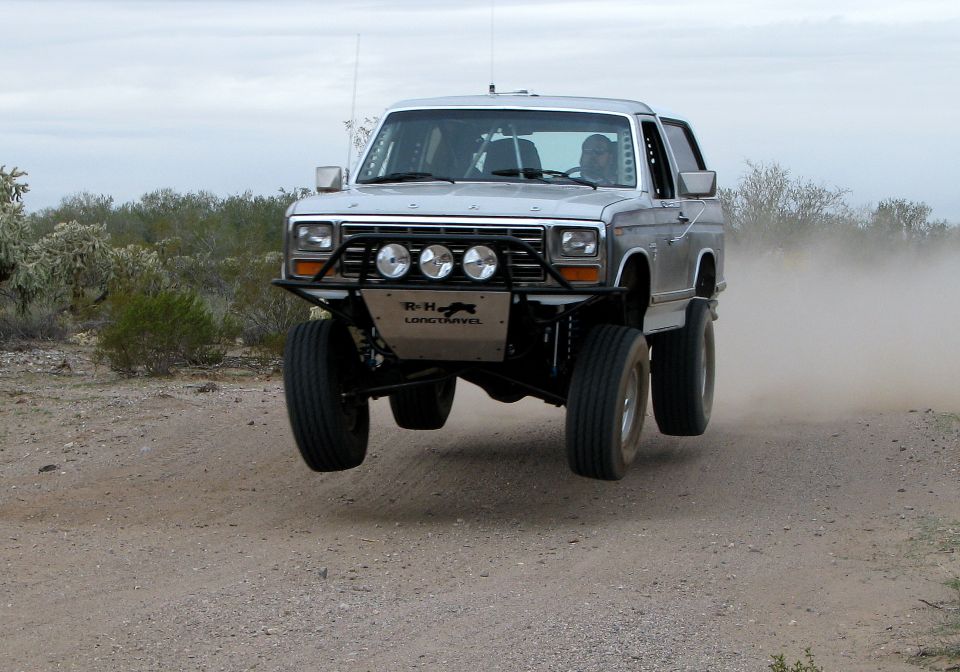 Silver Ford Bronco mid-air jump on a dirt road, desert landscape