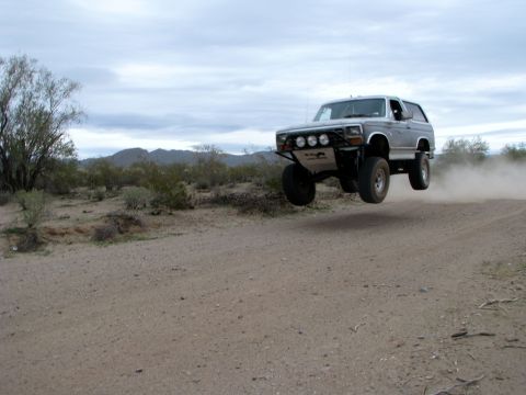 1980 Ford Bronco - A silver 1980 Ford Bronco takes flight over a dusty desert road.