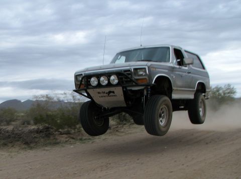 1980 Ford Bronco - A gray 1980 Ford Bronco is captured mid-air during a jump over a dirt track in a desert environment.