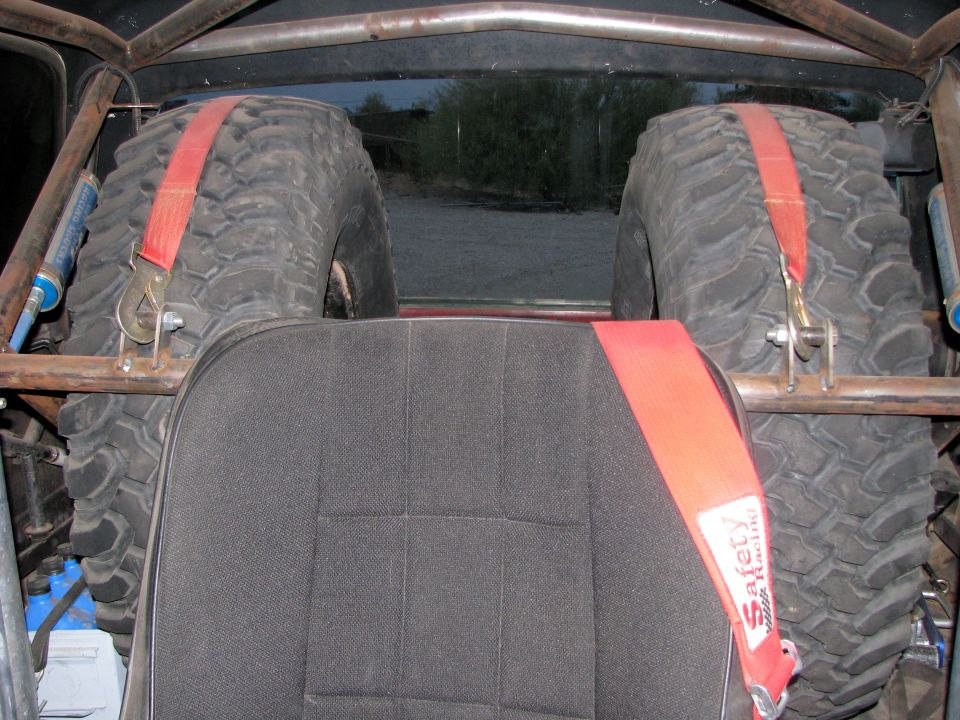 Interior view of a roll cage with two spare tires strapped in behind the seats. Red safety harness visible