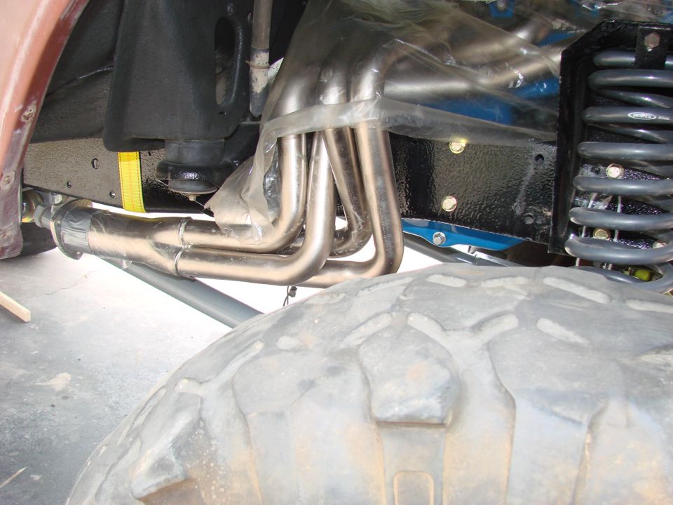 Close-up of shiny exhaust headers and suspension parts under a vehicle, with a large tire in the foreground