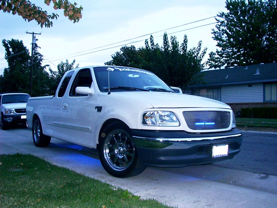 Lowered white Ford F-150 extended-cab pickup with billet grille, chrome wheels, and blue underglow in a driveway