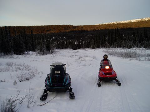 2000 Ford E350 - Two snowmobiles are parked on a snowy trail, with a dense forest and hills visible in the distance under a clear sky.