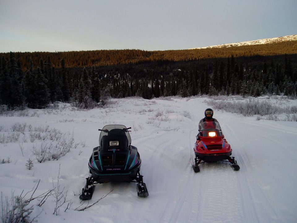 Two snowmobiles parked on a snow-covered trail with a forest and hills in the background