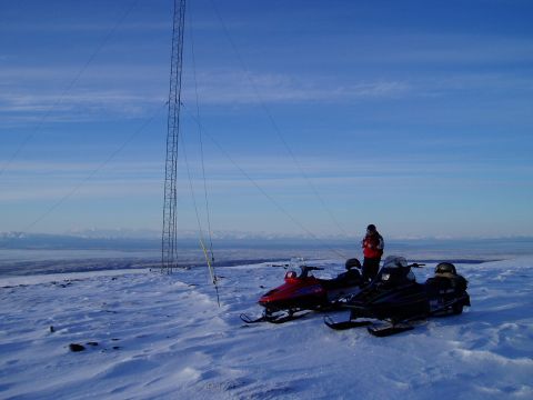 2000 Ford E350 - A person stands near two snowmobiles on a snow-covered mountain, with a large tower and distant mountains under a clear blue sky.