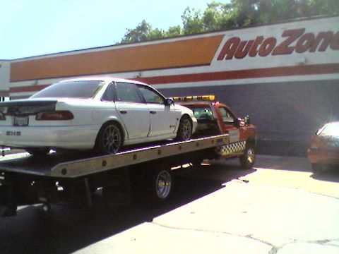 1993 Ford Taurus - A white 1993 Ford Taurus sedan is transported on a tow truck in front of an Auto Zone store.