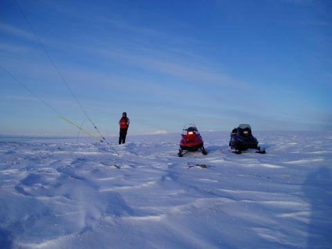 2000 Ford E350 - A person stands in a wide, open, snow-covered field with two snowmobiles nearby, under a bright blue sky.