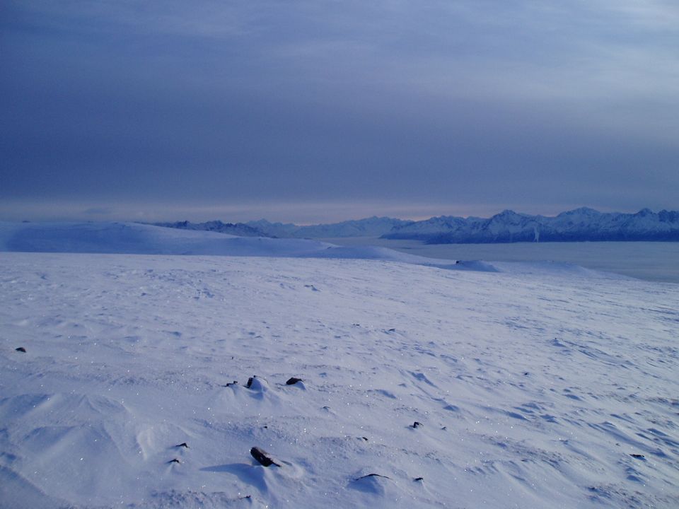 Snowy landscape with wind-swept snowdrifts and distant mountains under a cloudy sky. A small dark object, possibly a vehicle, is visible in the far distance