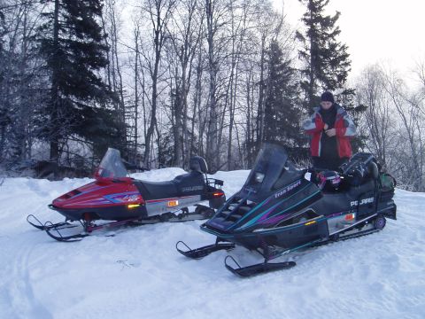 2000 Ford E350 - Two snowmobiles are parked in a snowy wooded area, with a person standing next to one of them.