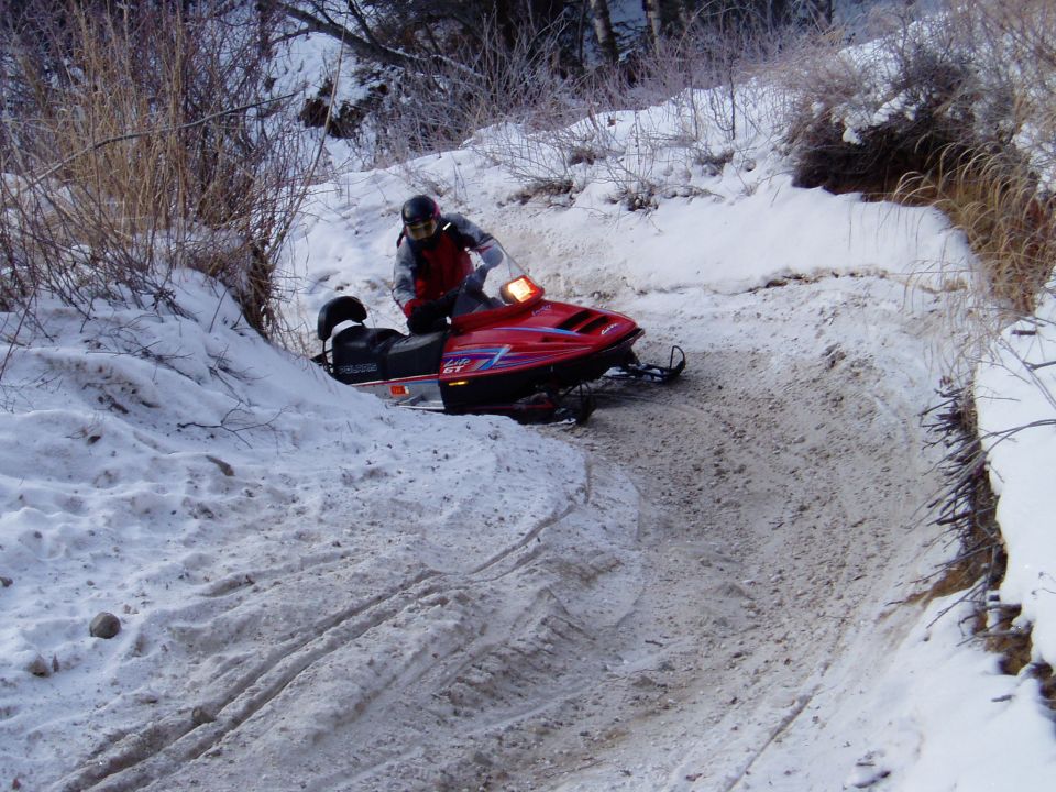 Red snowmobile with rider on a snowy trail, with trees and brush