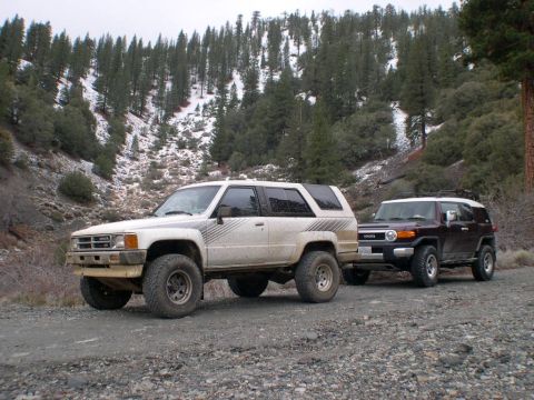 2007 Toyota FJ Cruiser - Two Toyota SUVs, one older and one newer, parked on a gravel road with a wooded, snow-dusted hillside behind them.