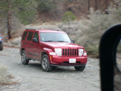 2007 Toyota FJ Cruiser - A red Jeep Liberty drives on a gravel road, with a portion of another vehicle visible to the left.