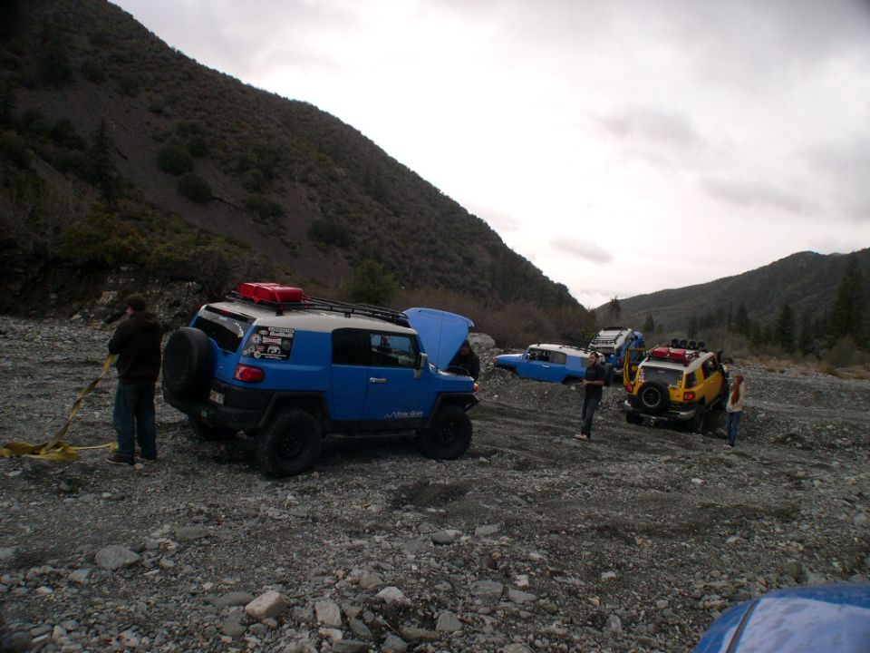 Several Toyota FJ Cruisers parked in a rocky, outdoor area with hills in the background. One has its hood up