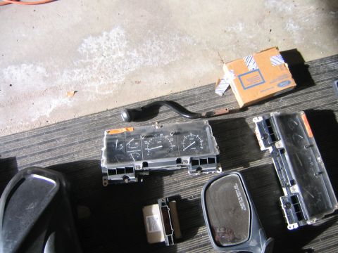 1994 Ford Bronco - Disassembled 1994 Ford Bronco parts, including the instrument cluster and side mirror, are laid out on a striped surface next to a Ford-bra…