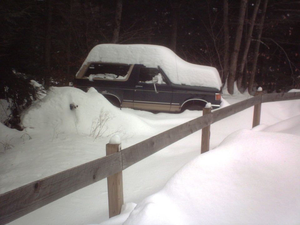 1980s Ford Bronco with a camper shell, partially buried in snow, parked next to a wooden fence