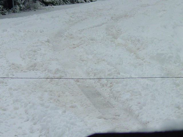 Snow-covered road with tire tracks and a dark vehicle bumper in the foreground