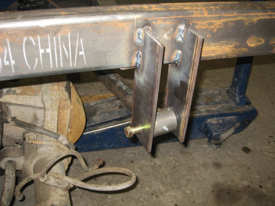 Custom fabricated bracket welded to a metal frame, with a bolt and bushing assembly. Vehicle axle and suspension components are visible to the left