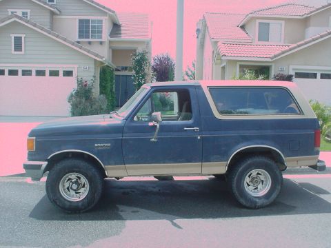 1986 Ford Bronco - Side profile of a blue 1986 Ford Bronco with tan trim, parked in front of houses.