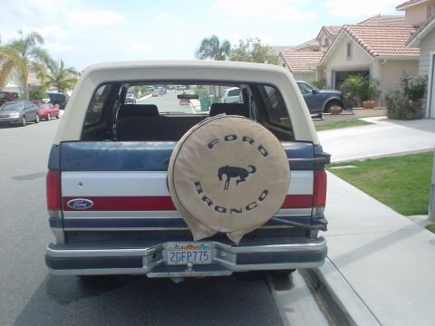 1986 Ford Bronco - A blue and white Ford Bronco is parked on a residential street, showing its rear with a spare tire cover.