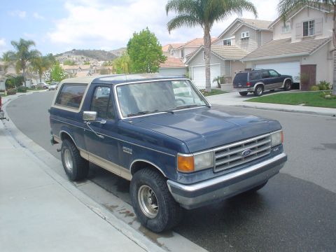 1986 Ford Bronco - This 1986 Ford Bronco, identified by its grille, is parked on a residential street with another SUV in the background.