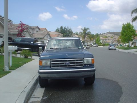 1986 Ford Bronco - Front view of a blue Ford pickup truck parked on a residential street. Another truck is visible in the driveway.