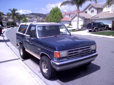 1986 Ford Bronco - A dark blue Ford Bronco is parked curbside on a street with houses and palm trees in the background.
