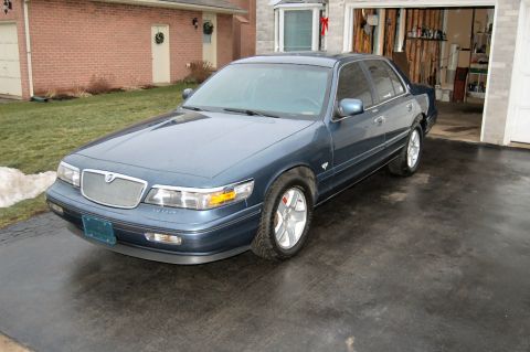 1985 Mercury Grand Marquis - A blue sedan is parked in a driveway with a garage in the background.