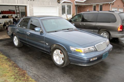 1985 Mercury Grand Marquis - A blue Mercury Grand Marquis is parked in a driveway, with a dark minivan behind it.