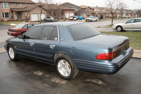 1985 Mercury Grand Marquis - A blue sedan is parked on a driveway in front of houses. Another red car is visible in the background.
