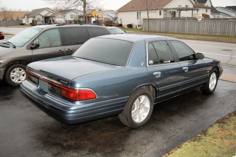 1985 Mercury Grand Marquis - A blue sedan is parked on a wet driveway next to a dark minivan.