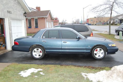 1985 Mercury Grand Marquis - A blue sedan is parked in a driveway next to a garage, with a van visible behind it.
