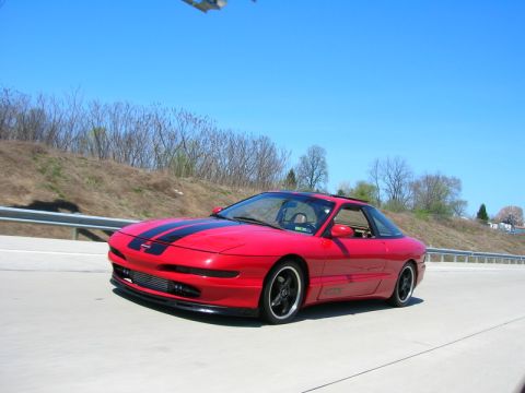 1994 Ford Probe - A red Ford Probe with dual black racing stripes cruises down the highway under a clear blue sky.