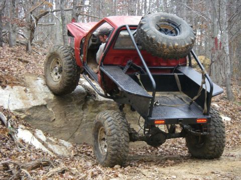 1978 Ford Bronco - A heavily modified red off-road vehicle navigates a rocky trail in a wooded area.