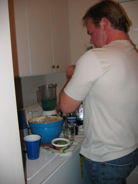 1985 Ford LTD Crown Victoria - A man prepares a drink in a kitchen setting, with a blender, blue bowl, and various bottles on the counter.