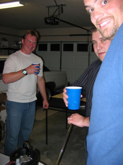 1985 Ford LTD Crown Victoria - Men gathered in a garage holding drinks, with garage doors visible behind them.