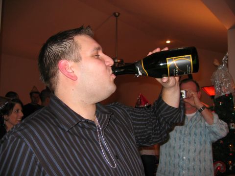 1985 Ford LTD Crown Victoria - A man drinks directly from a champagne bottle while others celebrate in the background, with one person capturing the moment with a camera.
