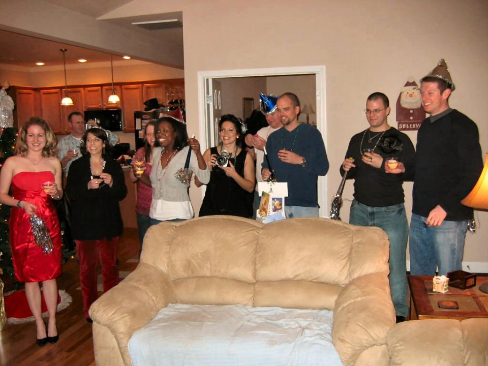 People in party hats and festive attire gathered in a living room, some holding drinks and cameras