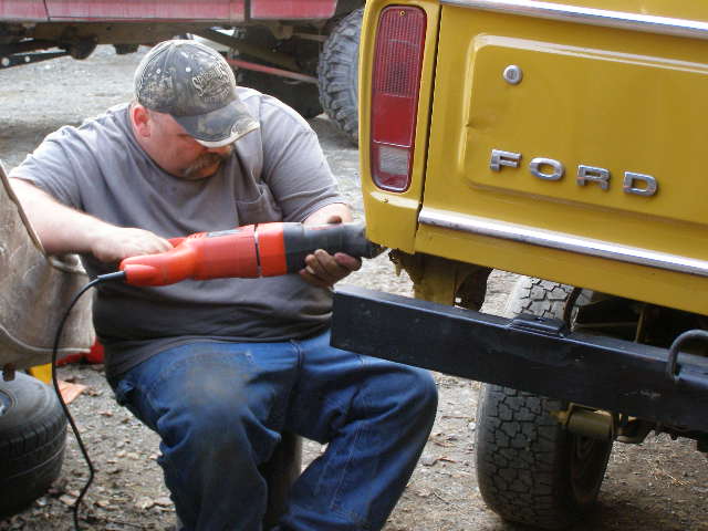 Man using a reciprocating saw on the rear panel of a yellow 1978 Ford Bronco. The word FORD is visible on the tailgate