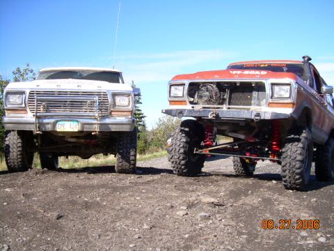 1978 Ford Bronco - Two heavily modified trucks, including a 1978 Ford Bronco, are parked side-by-side on a dirt road.