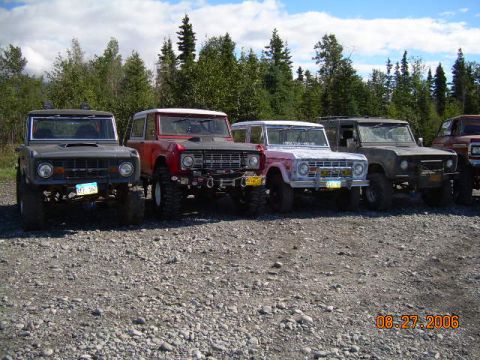 1978 Ford Bronco - A lineup of four Ford Broncos, including a gray, red, pink, and olive green model, are parked on a gravel lot with trees in the background.