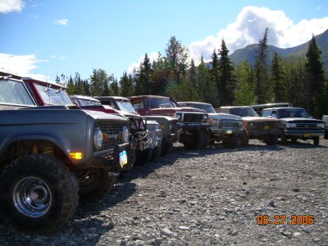 1978 Ford Bronco - A line of SUVs and trucks parked on a gravel lot with trees and mountains in the background.