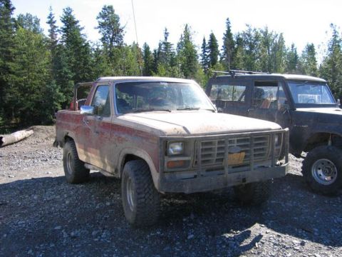 1978 Ford Bronco - Two SUVs, one a 1978 Ford Bronco, are parked on a gravel lot with trees in the background.