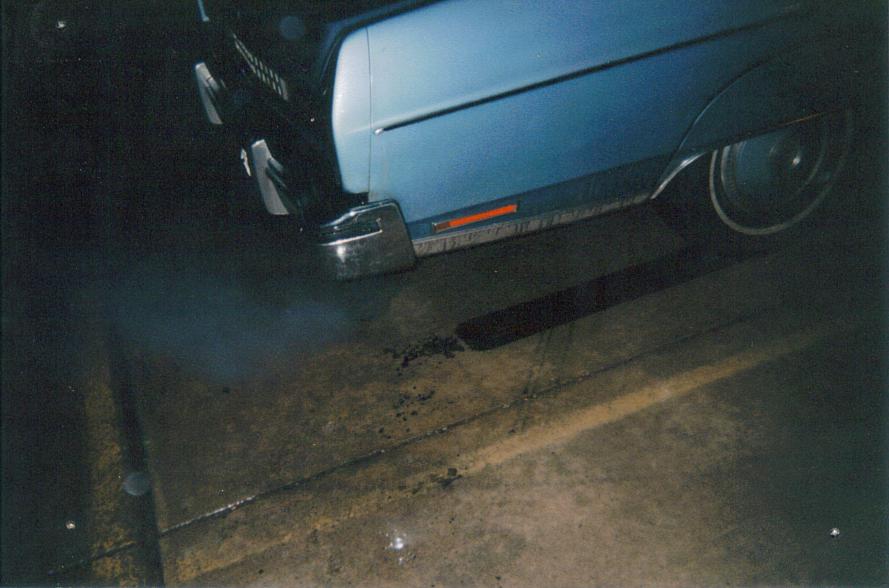 Front corner of a light blue full-size car at night with smoke near the pavement