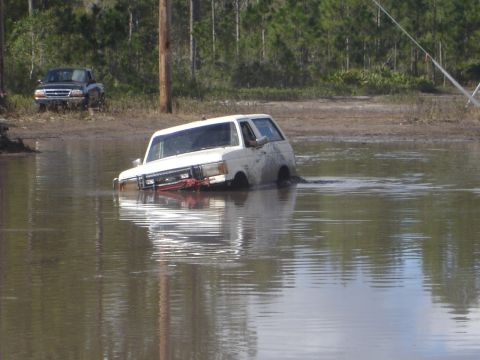 1989 Ford Bronco - A white SUV is partially submerged in muddy water, with another truck visible in the background.