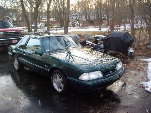 1991 Ford Mustang - A green 1991 Ford Mustang is parked on a wet driveway next to a red pickup truck.