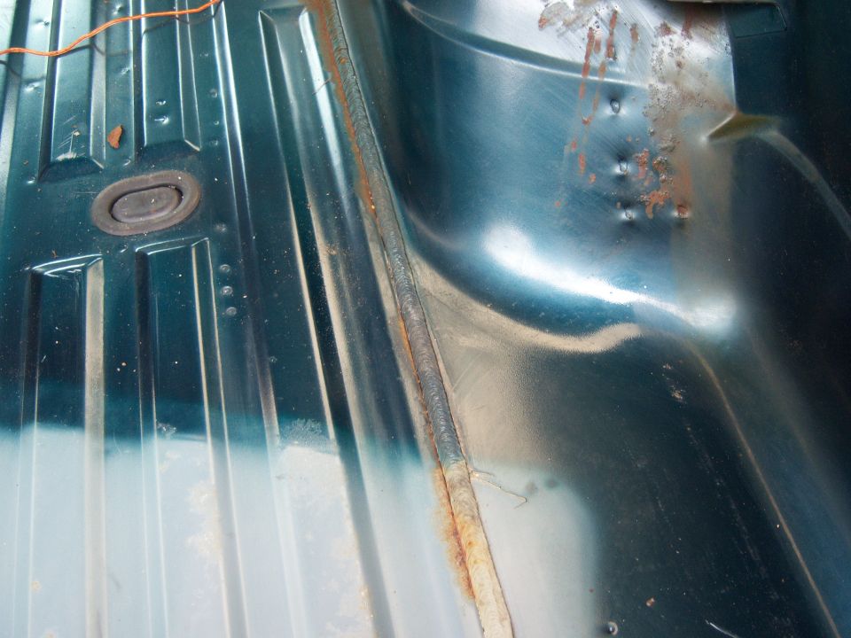 Interior floor of a vehicle showing ribbed metal panels and a rubber grommet. Some rust is visible