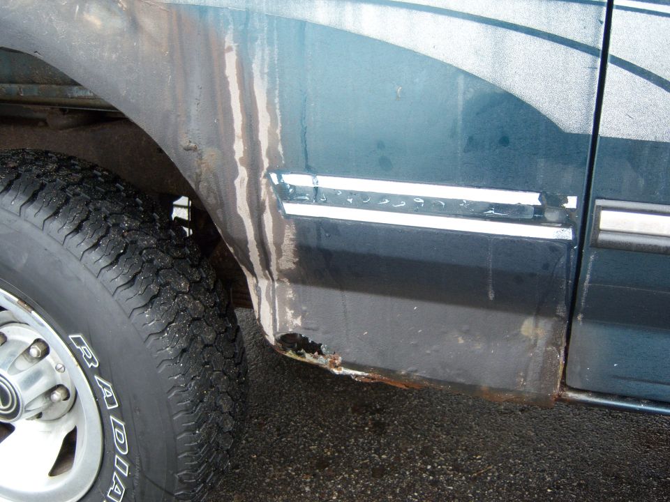 Close-up of a dark teal vehicle's lower rear quarter panel showing rust and water streaks near the wheel well