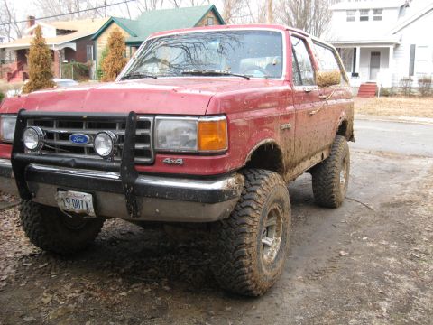 1988 Ford Bronco - This red Ford Bronco is covered in mud, suggesting it has been used off-road. It features a bull bar and large tires.