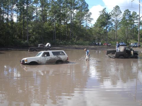 1989 Ford Bronco - Several vehicles are stuck in a muddy area, with trees and a cloudy sky in the background.